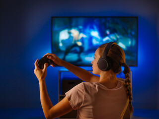 Canvas Print - A young girl with headphones and a joystick in her hands plays a video game in front of a large computer monitor. Game strategy, free time, hobbies, relaxation, rest.