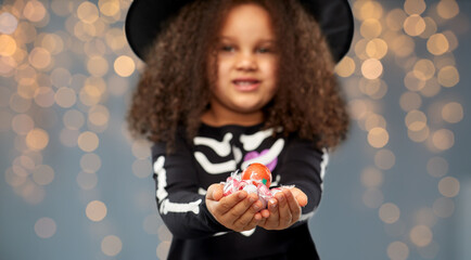 Wall Mural - halloween, holiday and childhood concept - african american girl in black costume and witch hat with candies trick-or-treating over lights on grey background