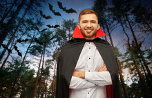 Holiday, Theme Party And People Concept - Happy Smiling Man In Halloween Costume Of Vampire And Dracula Cape With Arms Crossed Over Bats Flying In Dark Night Forest Background