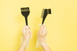 set for home or salon hair dyeing in the hands of a woman with gloves. Brushes and bowl for hair dye on yellow background