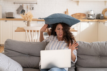 Wall Mural - Furious young woman try to concentrate on work as small disobedient kid hit her with pillow. Angry mother freelancer or distance office worker on lockdown at home with hyperactive preschooler child