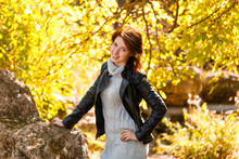 Happy Young Woman In Park On Sunny Autumn Day. Cheerful Beautiful Girl In Gray Sweater And In A Leather Jacket Outdoors On A Beautiful Autumn Day. Natural Light.