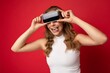 © Ivan Traimak - Portrait photo of funny young blonde woman wearing white t-shirt isolated on red background with copy space holding smartphone showing phone in hand with empty screen for cutout and showing tongue
