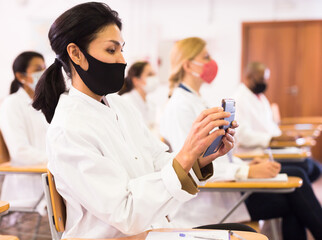 Wall Mural - Asian female doctor in protective face mask attending medical congress, using mobile phone to record lecturer speech. New normal during coronavirus pandemic