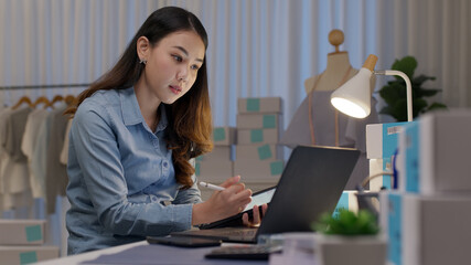 Wall Mural - Candid young asia freelance seller woman happy work on laptop notebook busy night at home office desk use digital tablet computer in SME online store social e-commerce sale page service web support.