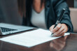 © Migue Suarez - Mujer joven firma en un papel un contrato de trabajo junto a su ordenador