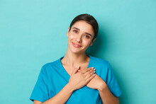 Coronavirus, Social Distancing And Health Concept. Close-up Of Smiling Attractive Female Doctor, Feeling Grateful, Holding Hands On Heart And Looking With Gratitude, Standing Over Blue Background