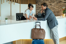 Businessman Using Digital Tablet For Checking In At Hotel.
Businessman With Luggage Standing At Hotel Reception And Filling In Registration Forms On Digital Tablet With Assistance Of Female Concierge.