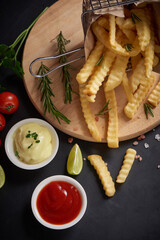 Wall Mural - Homemade baked potato fries with mayonnaise, tomato sauce and rosemary on wooden board. tasty french fries on cutting board, in brown paper bag on black stone table background, unhealthy food.