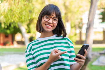 Wall Mural - Young brunette woman in the park using mobile phone and pointing it