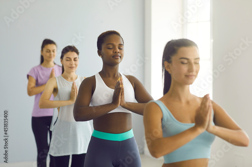 Group Meditation. Relaxed afro american young woman standing with folded arms in namaste gesture at group yoga class. Woman meditates among other caucasian women in a bright hall. Selective focus.