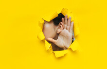 Close-up Of A Woman's Ear And Hand Through A Torn Hole In The Paper. Yellow Background, Copy Space. The Concept Of Eavesdropping, Espionage, Gossip And Tabloids.
