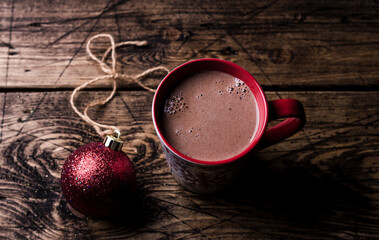 Delicious hot chocolate cocoa drink in red mug with christmas red ball and gift on wooden background, traditional winter drink, selective focus, top view, space, toned	