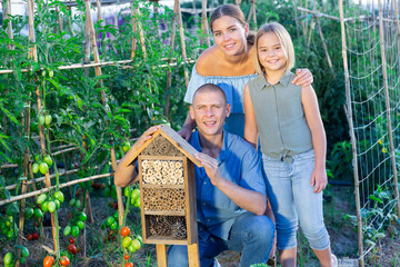 Wall Mural - Happy family next to wooden insect hotel in form of birdhouse between the beds with tomatoes. High quality photo