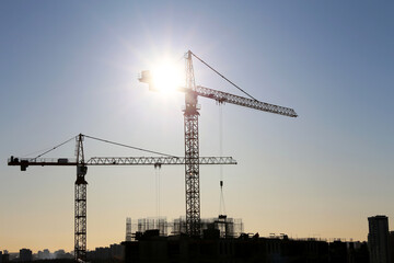 Wall Mural - Silhouettes of construction cranes on unfinished residential building against the sky and shining sun. Housing construction, apartment block in city