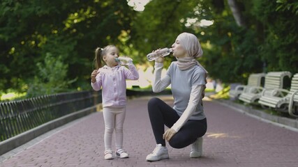 Wall Mural - Happy Muslim Arab mother looking at her smiling daughter holding bottle with water and drinking while resting after sport workout outdoors in the public park in the summer day