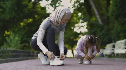 Wall Mural - Positive good-looking young mother in hijab, tying laces together with her little daughter, ready to do physical activity outdoors. Family, sport and recreation concept