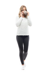 Wall Mural - Young woman in fluffy comfy sweater drinking coffee walking and talking on the phone. Full body portrait isolated on white background