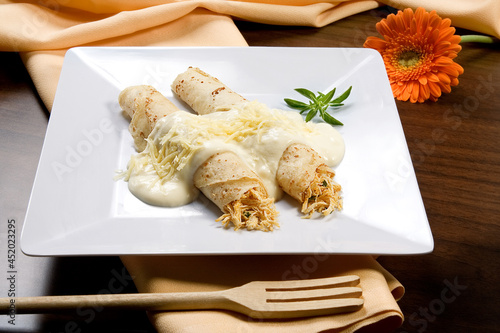 Panquecas com recheio de frango desfiado com molho bechamel e queijo parmesão, em prato branco no fundo de mesa decorada.