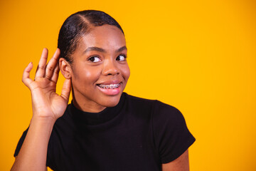 Gossip concept. Curious young afro woman holding hand near ear, trying to hear interesting information, cheerful korean female eavesdropping on yellow studio background, panorama with copy space.