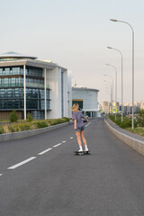 Female skateboarder skating on empty road. Blonde woman in t-shirt and jeans shorts riding longboard, back view. Active caucasian young girl has fun on skate. Urban lifestyle and extreme hobby concept