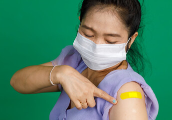 Wall Mural - Studio close up shot of Asian female patient with face mask look at camera pointing at yellow bandage plaster after receive coronavirus covid 19 vaccination from doctor at clinic on green background