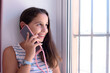 © scar - Brunette girl, sitting by the window of her house, smiles and looks at the street, while talking on her cell phone.