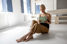 Stylish Woman Using A Digital Tablet While Sitting And Relaxing On The Floor At Modern Studio Apartment. Digital Lifestyle And Leisure Time At Cozy Home