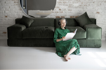 Wall Mural - Stylish woman in green dress reading book and listening to the music, while sitting on the floor near the couch at home. Leisure time and comfort living at home