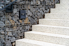 Granite Staircase With Fence Made Of Stones In Metal Grid And Black Lamp Built Into Wall To Illuminate Stairs At Night. Modern Concrete Staircase With Gabion Fence.