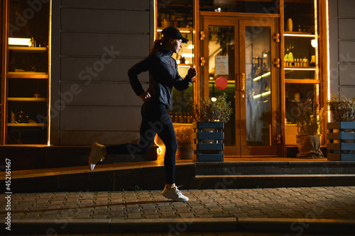 Young good-looking sportswoman is running in night city at streets, past around cafe, redhead lady in black outfit is engaged in workout cardio training, copy space. adult woman outdoors, fitness