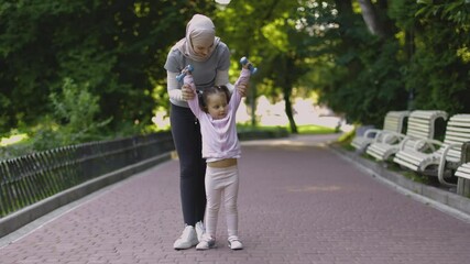 Wall Mural - Positive Muslim mother and daughter with barbells in the summer park. Sporty fit Arab woman showing her little kid working out with dumbbells, making raises up