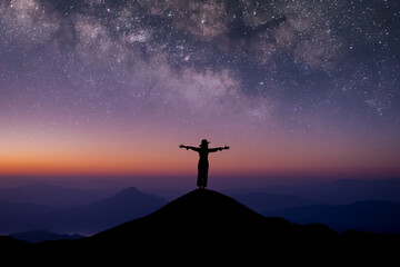 Wall Mural - Silhouette of young female traveler wearing hat standing and open arms watched the beautiful night sky, star and milky way alone on top of the mountain.