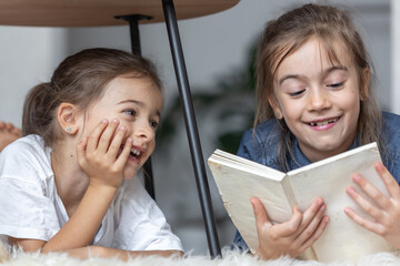 Poster - Two little sisters are reading a book lying on the floor on a cozy blanket.