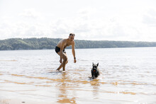 A Cheerful Young Dog Of The Scotch Terrier Breed Runs Along The Beach Against The Background Of The River. A Young Tall Caucasian Man Is Playing With A Dog. The Concept Of Outdoor Recreation. 