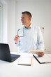 © epiximages - business man with blue shirt and black glasses is standing behind standing table and is working with his tablet and a black laptop in a modern office