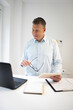 © epiximages - business man with blue shirt and black glasses is standing behind standing table and is working with his tablet and a black laptop in a modern office