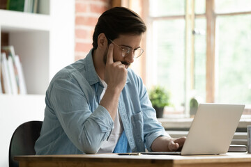 Serious man sit at desk look at laptop screen read e-mail ponder over received information, working remotely, learn new professional business application use modern tech develop online project concept