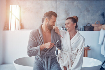 Wall Mural - Beautiful young couple in bathrobes smiling and cleaning teeth while doing morning routine