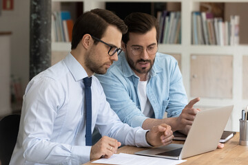 Two serious, confident male office colleagues review collaborative corporate task, discussing on-line business common project using computer. Teamwork, mentoring, partnership, commercial offer concept