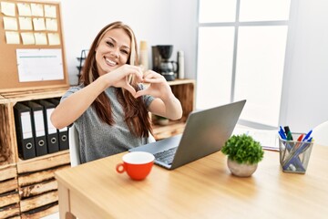 Poster - Young brunette woman working at the office with laptop smiling in love doing heart symbol shape with hands. romantic concept.