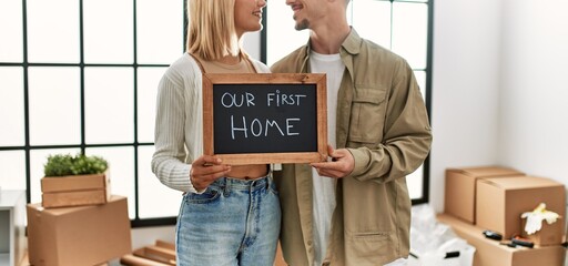 Poster - Young caucasian couple smiling happy holding blackboard with our first home message at new house