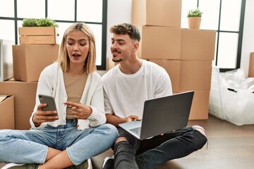 Wall Mural - Young caucasian couple smiling happy using laptop and smartphone sitting at new home.