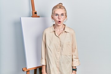 Poster - Beautiful caucasian woman with blond hair standing by painter easel stand scared and amazed with open mouth for surprise, disbelief face