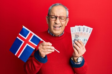 Poster - Handsome senior man with grey hair holding iceland flag and icelandic krona banknotes winking looking at the camera with sexy expression, cheerful and happy face.