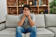 © Krakenimages.com - Young hispanic man wearing casual clothes sitting on the sofa at home shocked covering mouth with hands for mistake. secret concept.