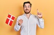Handsome caucasian man with beard holding denmark flag smiling with an idea or question pointing finger with happy face, number one