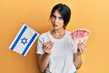 Sticker - Young caucasian woman holding israel flag and shekels banknotes in shock face, looking skeptical and sarcastic, surprised with open mouth