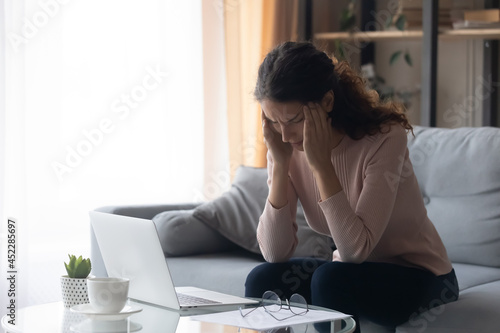 Young woman sit on sofa near laptop takes off glasses touch temples suffers from migraine headache high blood pressure, having eyes train after long time work at home. Blurry vision, bad news concept