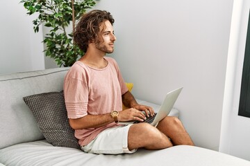 Poster - Young hispanic man sitting on the sofa at home using laptop looking to side, relax profile pose with natural face and confident smile.